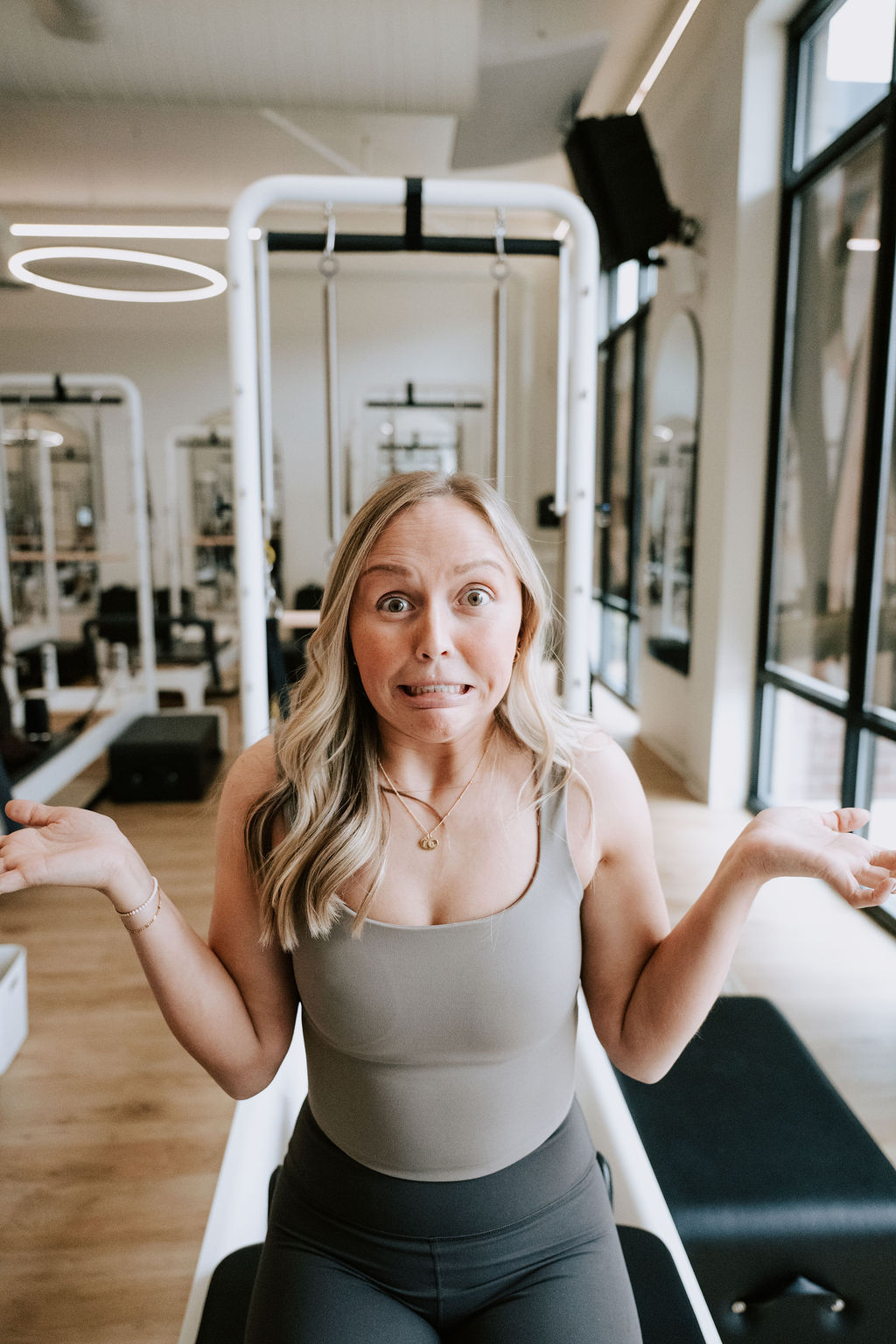 A client in a taupe tank top sits on a Reformer with a playful, questioning expression in a bright Core Collective studio